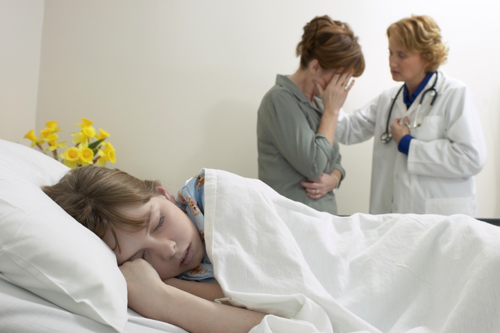 A young child laying in bed with a mother and doctor in the background.