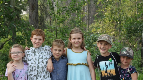 six children standing in a line with arms around one another's shoulders outdoors