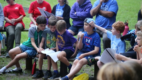 a group of people outdoors around a campfire, with a group of boys gathered around a song sheet